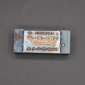 May include: A rectangular metal printing block featuring a copper-colored raised surface with the text A PRODUCT OF Westing MADE IN U. S. A. The block has a dark grey metallic base with visible rivets and a small brown blemish on the left side.