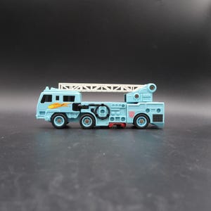 Puede incluir: Un camión de bomberos de juguete de color azul claro con una escalera blanca en la parte superior. El vehículo cuenta con ventanas negras, un gráfico de llama amarilla en el lateral, una rueda de repuesto negra y un símbolo Autobot rojo.