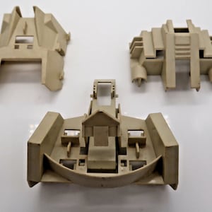 May include: Three tan plastic toy parts are arranged on a white surface. The parts have a geometric design with rectangular and curved shapes. The parts appear to be components of a larger toy or model.