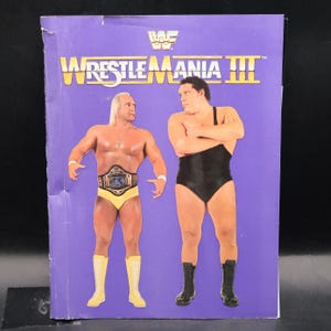 May include: A vintage purple WWF WrestleMania III promotional poster featuring Hulk Hogan and Andre the Giant. The poster has the WWF logo at the top, with the event title in yellow and white. Hogan is wearing yellow trunks and boots, and Andre is in a black singlet.