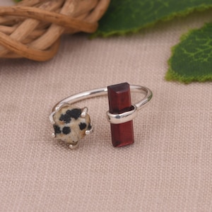 May include: A silver ring with a red gemstone and a white and black speckled gemstone. The ring is open and adjustable.