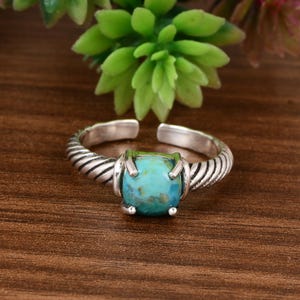 May include: A silver-toned ring with a turquoise stone. The ring has a twisted band design and an open adjustable style. The turquoise stone is square-shaped and set in a bezel setting. The ring is displayed on a wooden surface.
