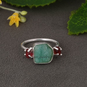May include: A silver ring with a large green gemstone and two smaller red gemstones. The ring is set on a dark gray background.