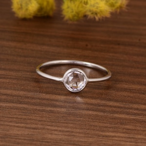 Op de afbeelding: Een zilveren ring met een heldere, ronde edelsteen in het midden. De ringband is dun en gepolijst, en de edelsteen is facetgeslepen en reflecteert licht. De ring wordt getoond op een houten oppervlak.