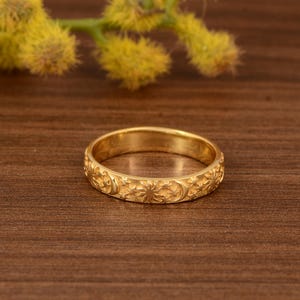 Könnte beinhalten: Ein goldfarbener Ring mit einem detaillierten floralen und himmlischen Muster. Der Ring weist geprägte Sonnen, Monde und florale Designs auf. Der Ring liegt auf einer Holzoberfläche mit gelben Blumen im Hintergrund.