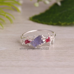May include: A silver ring featuring a rough-cut, light purple gemstone flanked by two red gemstones. The ring's design incorporates branch-like elements. The text "silvergemjewelry2022" is visible on the ring.