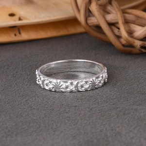 May include: A silver-colored metal ring featuring a repeating embossed pattern of suns, crescent moons, and small stars around the entire outer band. The ring has a polished finish and a consistent width, showcasing celestial motifs in a continuous design.
