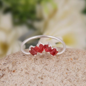 May include: A silver ring with a row of five red gemstone accents. The ring is set on a light brown stone surface.