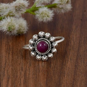 May include: A silver ring featuring a central round cabochon-cut deep magenta stone. The stone is surrounded by a twisted rope-style metal border and an outer halo of small, smooth silver spheres arranged in a circular floral-like pattern.