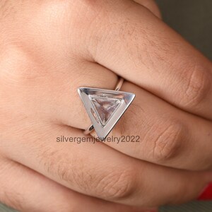 Puede incluir: Un anillo de plata con un engaste triangular que presenta una piedra preciosa transparente. El anillo est&aacute; en una mano.