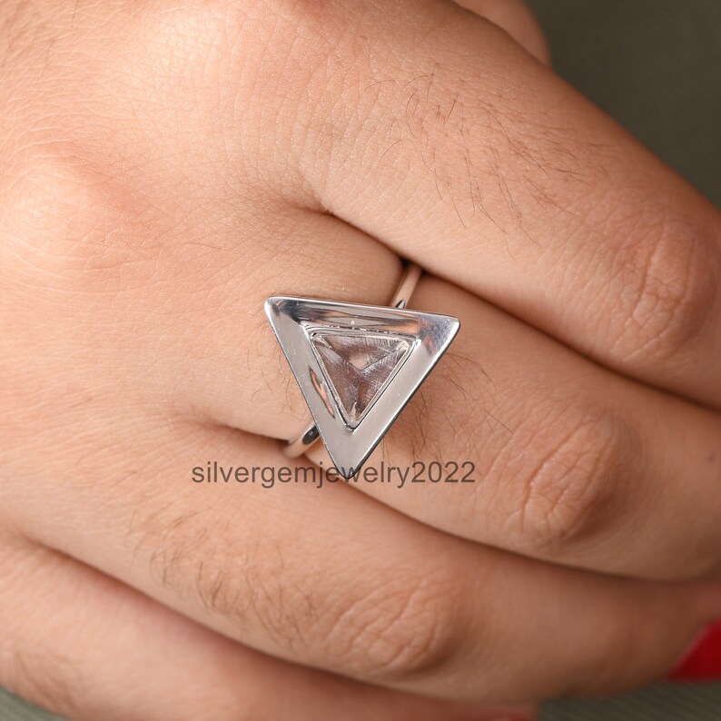 Puede incluir: Un anillo de plata con un engaste triangular que presenta una piedra preciosa transparente. El anillo est&aacute; en una mano.