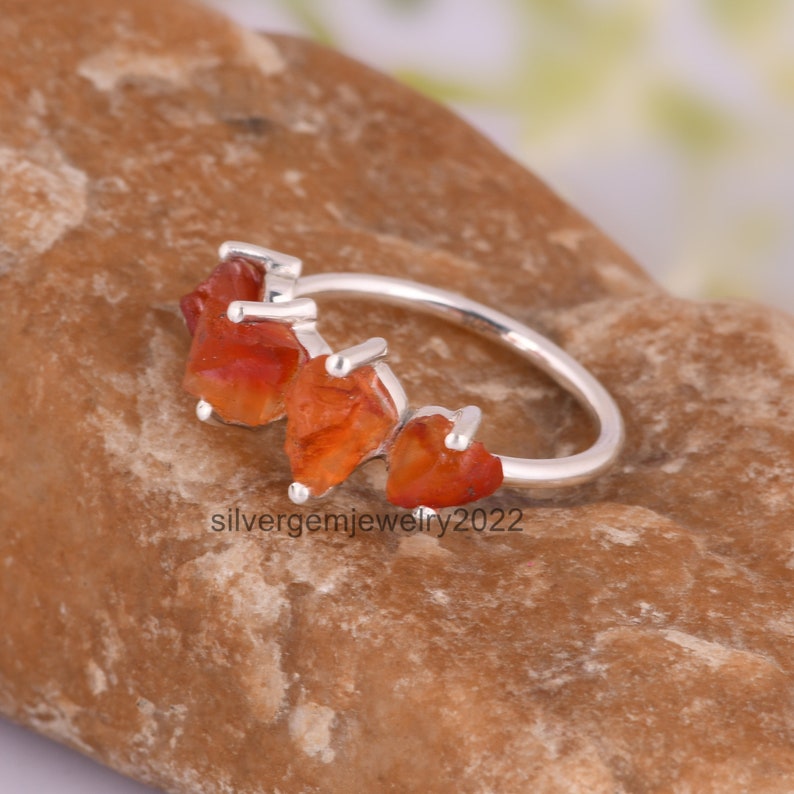May include: A silver ring with four orange carnelian stones set in a row. The stones are irregularly shaped and have a rough, natural look. The ring is a delicate and unique piece of jewelry.