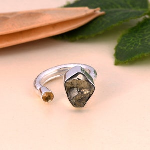 Puede incluir: Un anillo de plata con una piedra grande, rugosa, de color gris-marrón y una piedra más pequeña, redonda, de color amarillo. El anillo está abierto y tiene un acabado texturizado.