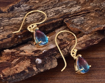 May include: A pair of gold-colored drop earrings with teardrop-shaped, multi-colored gemstones. The gemstones transition from blue to purple. The earrings are displayed on a wooden surface.