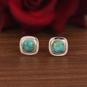 May include: Two square turquoise stud earrings with a silver border. The earrings are displayed on a wooden surface, with a blurred red flower in the background. The turquoise stones have a mottled appearance.