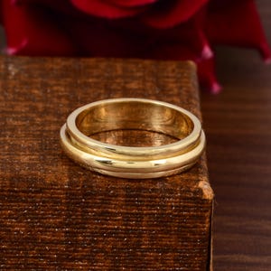 May include: A gold-colored ring with a double-band design. The ring is set against a dark brown surface, with a blurred red flower in the background. The ring's bands have a smooth, polished finish.