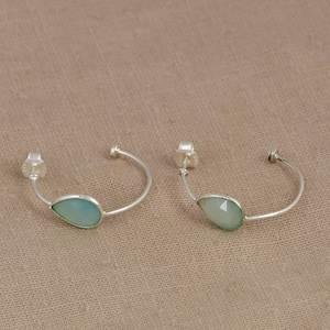 Peut inclure: Une paire de créoles en argent, chacune ornée d'une pierre précieuse en forme de goutte d'un bleu-vert pâle, fixée sur le fin anneau en métal poli. Les boucles d'oreilles présentent un design ouvert avec une petite tige de fermeture.