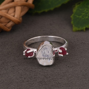 May include: A silver ring featuring a large, rough-cut moonstone and two smaller red ruby stones on either side. The ring is set in a simple band.