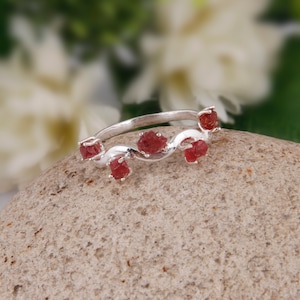 以下が含まれることがあります： ねじれたバンドと5つの赤い宝石が付いたシルバーリング。リングは灰色の石の背景に置かれています。