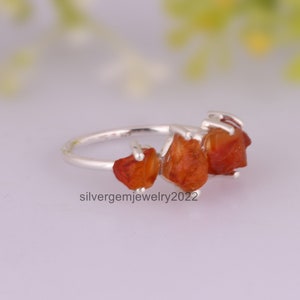 May include: A silver ring with three orange carnelian stones set in a raw, natural style. The stones are irregularly shaped and have a rough, unpolished texture.