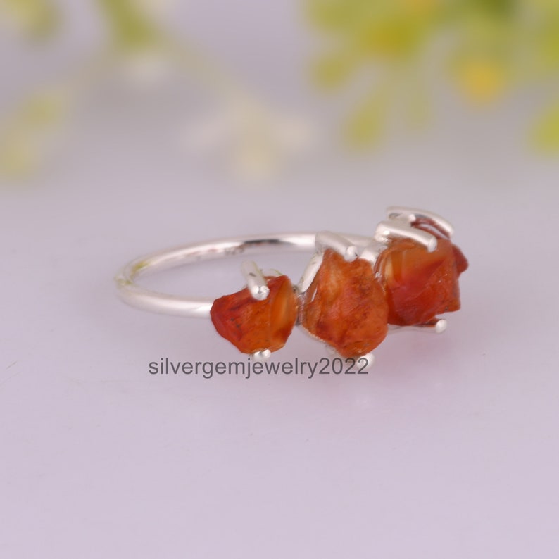 May include: A silver ring with three orange carnelian stones set in a raw, natural style. The stones are irregularly shaped and have a rough, unpolished texture.