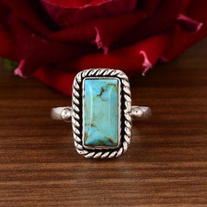 May include: A silver ring with a rectangular turquoise stone. The turquoise is a vibrant blue-green color with brown veining. The ring has a braided rope design around the stone and a simple band.
