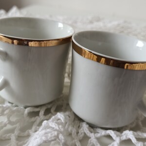 Puede incluir: Dos tazas de café de cerámica blanca con ribete dorado. Las tazas están sobre una superficie de crochet blanco.