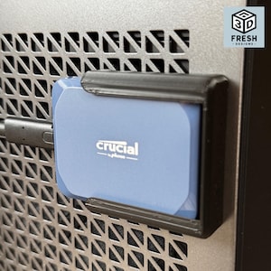 May include: A blue Crucial by Micron portable solid state drive secured in a black 3D-printed plastic mounting bracket. The drive is connected by a black cable and attached to a dark gray metal mesh surface. A logo reads 3D FRESH DESIGNS.