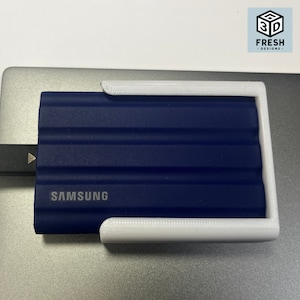 May include: A navy blue Samsung external hard drive connected to a laptop with a USB-C cable. The hard drive is held in place by a white 3D-printed holder. The Fresh Designs logo is visible in the top right corner.