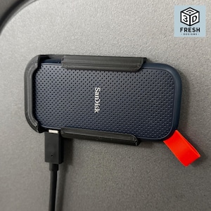 May include: A dark blue SanDisk portable solid state drive held in a black plastic mount. The drive features a perforated surface texture and a bright orange rubber loop on the side. A black USB-C cable is plugged into the bottom of the device.