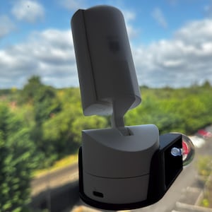 May include: A white cylindrical security camera mounted on a black plastic bracket with a clear suction cup attached to a glass window. The camera features a vertical hinge, a small lens area, and a visible charging port at the base of the unit.