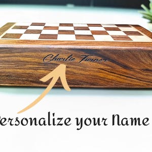 K&ouml;nnte beinhalten: Ein Schachspiel aus Holz mit Schachbrettmuster und dunkelbraunem Finish. Der Name "Charlie Turner" ist auf der Seite eingraviert. Der Text "Personalisiere deinen Namen" steht darunter.