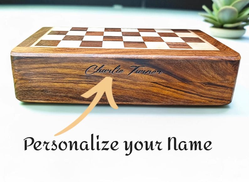K&ouml;nnte beinhalten: Ein Schachspiel aus Holz mit Schachbrettmuster und dunkelbraunem Finish. Der Name "Charlie Turner" ist auf der Seite eingraviert. Der Text "Personalisiere deinen Namen" steht darunter.