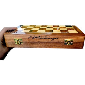 K&ouml;nnte beinhalten: Ein Schachspiel aus Holz mit einem Schachbrettmuster in hellen und dunklen Holzt&ouml;nen. Die Box ist braun mit goldfarbenen Verschl&uuml;ssen und der Name "Montemayor" ist auf der Vorderseite geschrieben.