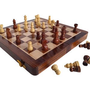 K&ouml;nnte beinhalten: Ein Schachspiel aus Holz mit einem dunkelbraunen Rahmen und einer hellen Holzspielfl&auml;che. Die Schachfiguren sind auf dem Brett platziert, wobei einige Figuren umgesto&szlig;en sind. Das Set ist ge&ouml;ffnet und spielbereit.
