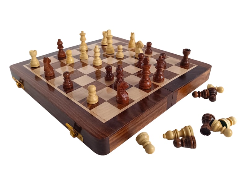 K&ouml;nnte beinhalten: Ein Schachspiel aus Holz mit einem dunkelbraunen Rahmen und einer hellen Holzspielfl&auml;che. Die Schachfiguren sind auf dem Brett platziert, wobei einige Figuren umgesto&szlig;en sind. Das Set ist ge&ouml;ffnet und spielbereit.