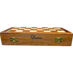K&ouml;nnte beinhalten: Ein Schachspiel aus Holz mit einem hellen und dunklen Schachbrett. Die Holzbox hat goldfarbene Verschl&uuml;sse und der Name "Louisa" ist auf der Vorderseite geschrieben. Das Schachspiel ist geschlossen und bereit f&uuml;r die Aufbewahrung oder den Transport.