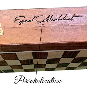 K&ouml;nnte beinhalten: Ein Schachspiel aus Holz mit personalisierter Inschrift. Die Oberseite der Box ist hellbraun mit einem dunkelbraunen Rand. Das Schachbrett ist ein kariertes Muster aus hellem und dunklem Holz. Die Box hat einen goldenen Verschluss.