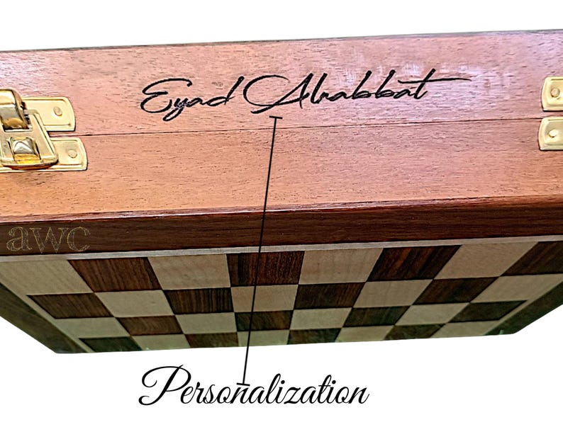 K&ouml;nnte beinhalten: Ein Schachspiel aus Holz mit personalisierter Inschrift. Die Oberseite der Box ist hellbraun mit einem dunkelbraunen Rand. Das Schachbrett ist ein kariertes Muster aus hellem und dunklem Holz. Die Box hat einen goldenen Verschluss.