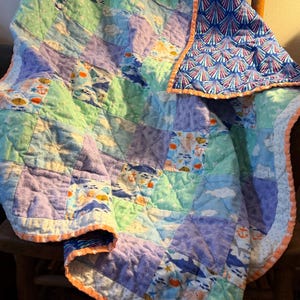 May include: A patchwork quilt featuring alternating squares of textured lavender, light green, and white fabric printed with ocean motifs including sharks, anchors, and seashells. The quilt has an orange trim and a blue shell-patterned backing.