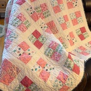 May include: Handmade quilt featuring a patchwork design with squares of various pink and white patterned fabrics. The quilt has a white background and is draped over a dark surface. The quilt is a cozy home decor item.