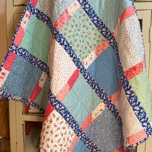 May include: A colorful patchwork quilt with a variety of patterns and colors. The quilt features squares of light blue, pink, white, and green fabrics, bordered by a dark blue floral print. The quilt is likely handmade.