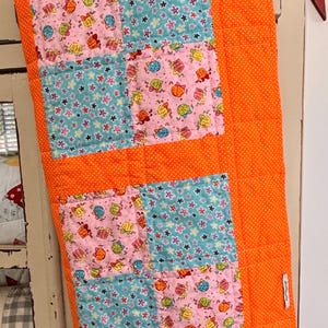 May include: A patchwork quilt with a bright orange background and squares of pink, blue, and white fabric with a floral and gift box pattern. The quilt has a small white tag with the word "Handmade" on it.