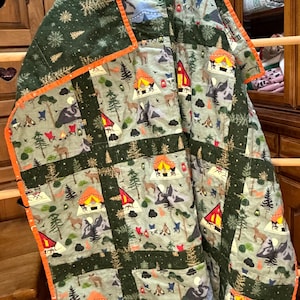 May include: A patchwork quilt featuring a repeating camping-themed print with orange, yellow, and red tents, brown deer, and dark green pine trees on a light gray background, finished with a bright orange fabric binding along the edges.