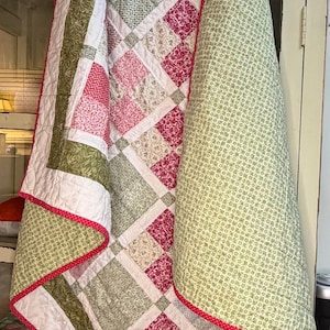 May include: A handmade quilt with a patchwork design. The quilt features squares of green, pink, and white fabric, bordered by a green and white floral pattern. The edges are finished with a red dotted trim. A cozy home decor item.