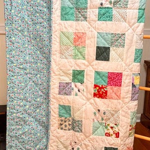 May include: A handmade quilt with a patchwork design. The quilt features squares of various patterns and colors, including pink, green, and floral prints, set against a cream background. The reverse side is a light blue fabric with a floral pattern.