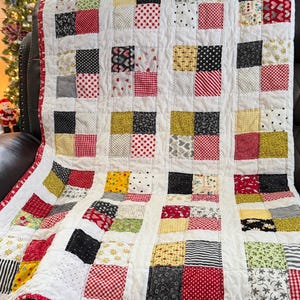 May include: A handmade patchwork quilt with a white background and a grid pattern of colorful squares. The squares feature various patterns in red, black, yellow, and white, including polka dots, stripes, and floral designs. The quilt is trimmed with a red border.