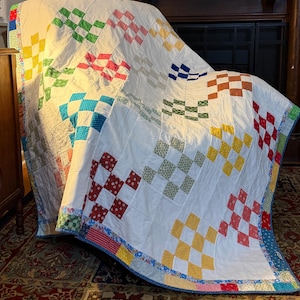 May include: A handmade quilt with a white background and colorful checkerboard patterns in shades of red, green, blue, and yellow. The quilt is draped over a dark surface, showcasing its patchwork design and detailed stitching.