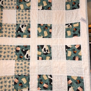 May include: A white quilt with a patchwork design featuring various farm animal prints. The quilt is made up of squares with different animal patterns, including cows, pigs, chickens, and sheep. The quilt has a white background with a subtle blue and white polka dot pattern.