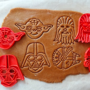 Op de afbeelding: Rode plastic koekjesvormen in de vorm van Star Wars-personages, waaronder Yoda, Chewbacca, Darth Vader en de Millennium Falcon. De vormen liggen op een bruin gingerbread-deeg.